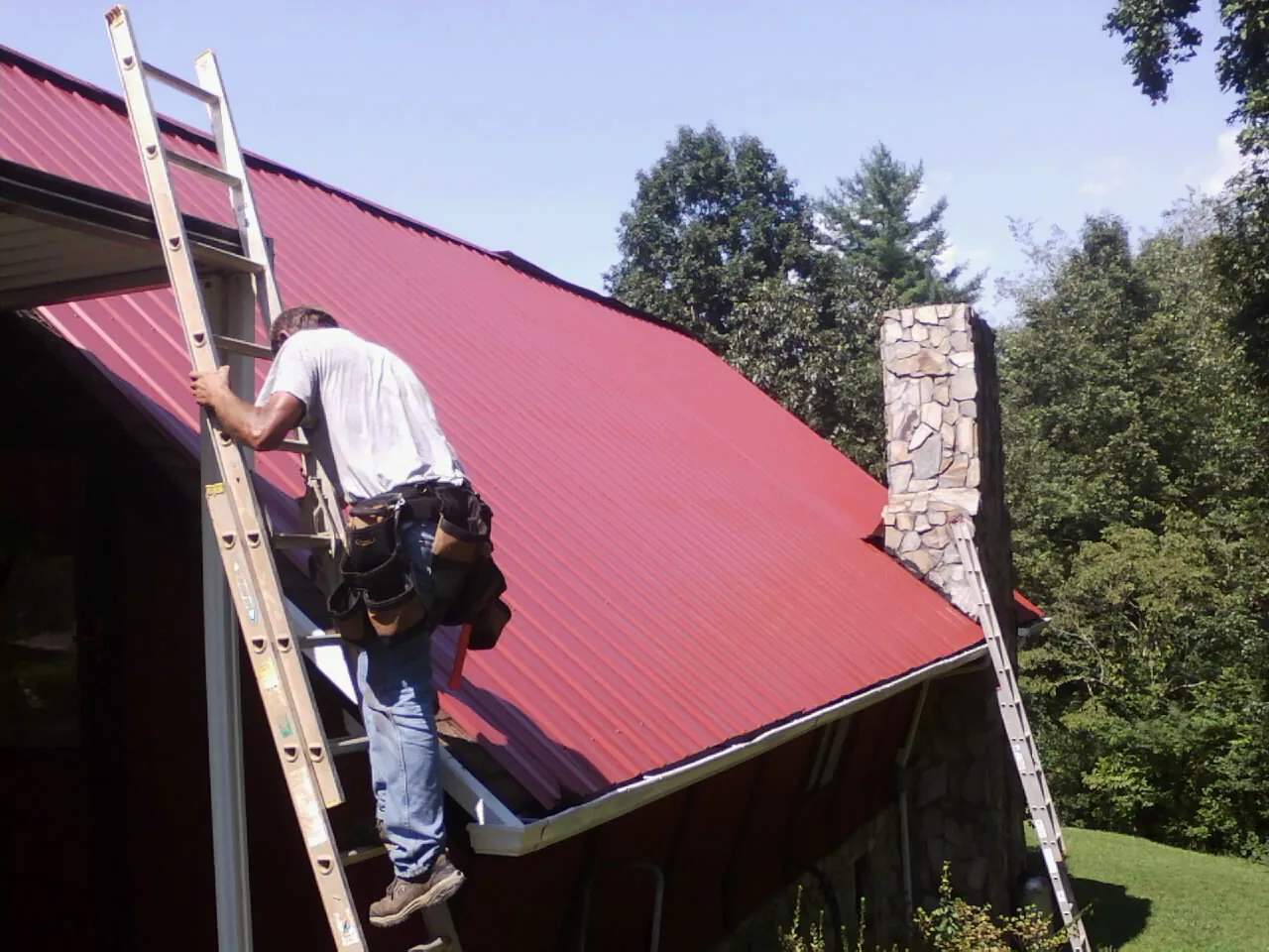 Roofer on a ladder beside a red metal roof and stone chimney — Soffit & Fascia Repair by Roofing Brothers in Teaneck